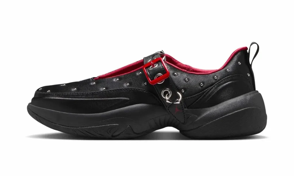 Side profile of a black slip-on shoe featuring a sculpted oversized midsole, satin-like upper with metallic studs, an adjustable black strap with a glossy red buckle, silver ring hardware, and a deep red interior lining