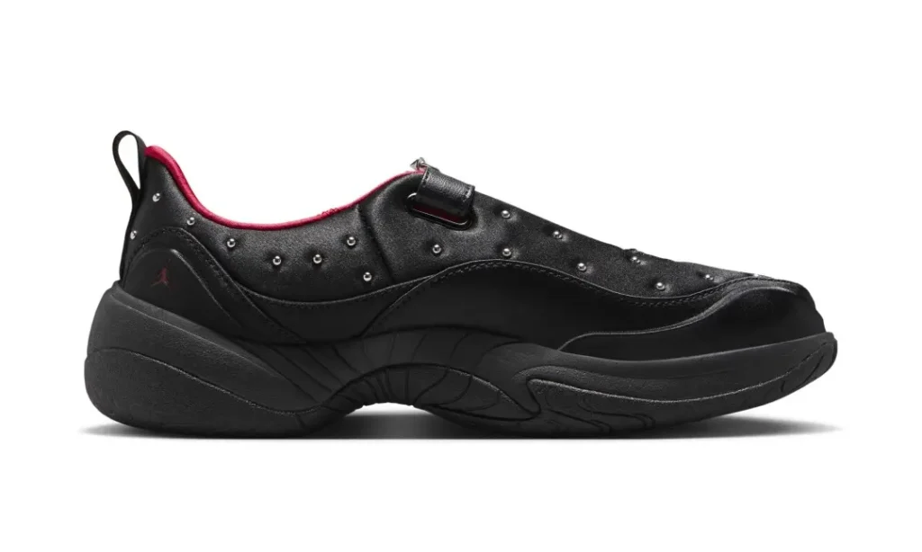 Medial side view of the black slip-on shoe highlighting the streamlined silhouette, sculpted foam sole, studded upper, red interior lining, heel pull tab, and subtle Jumpman logo near the heel