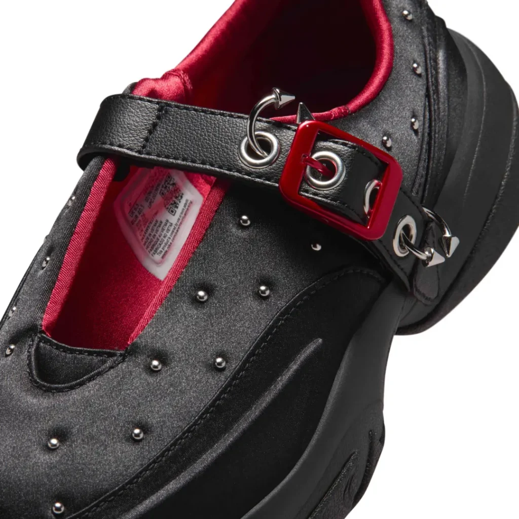 Close-up of the adjustable strap system featuring a glossy red square buckle, silver grommets, rings, pointed hardware, black leather strap, and satin black upper with metallic studs against a rich red lining