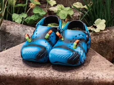 Pair of blue and black Brain Dead x Crocs clogs displayed on a weathered stone ledge beside a fountain, featuring swirling molded graphics, ventilation ports, adjustable heel straps, and translucent amber-and-green Jibbitz charms on the toe box