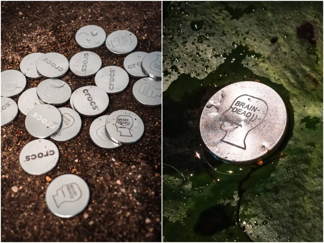 A close-up collage of silver Crocs Jibbitz charms scattered across wet pavement beside a fountain, including several classic Crocs logo discs and a limited BRAIN DEAD profile-head emblem resting in shallow green-tinted water.
