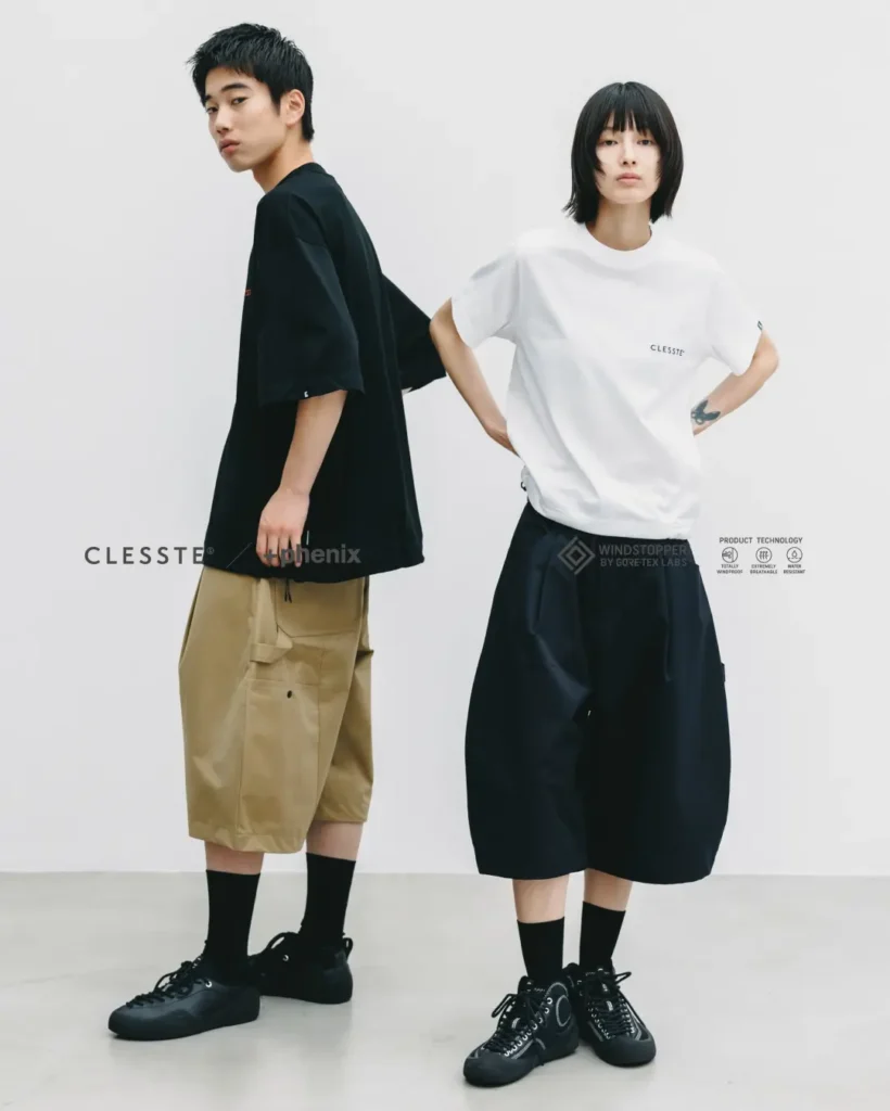 Two models wearing the CLESSTE × +phenix collection, styling oversized black and khaki WINDSTOPPER utility half pants with relaxed-fit T-shirts and monochrome footwear