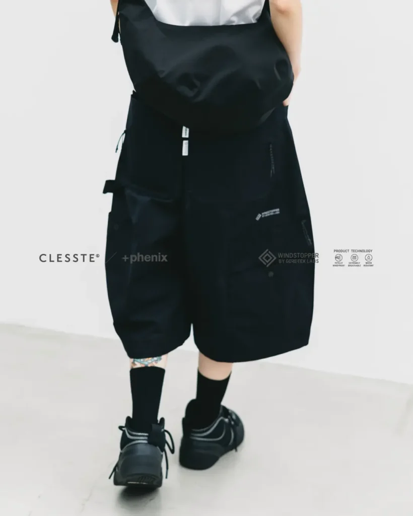 Rear view of a model wearing oversized black technical balloon shorts with large utility pockets, a black crossbody bag, black crew socks, and black low-top shoes against a minimalist white studio backdrop