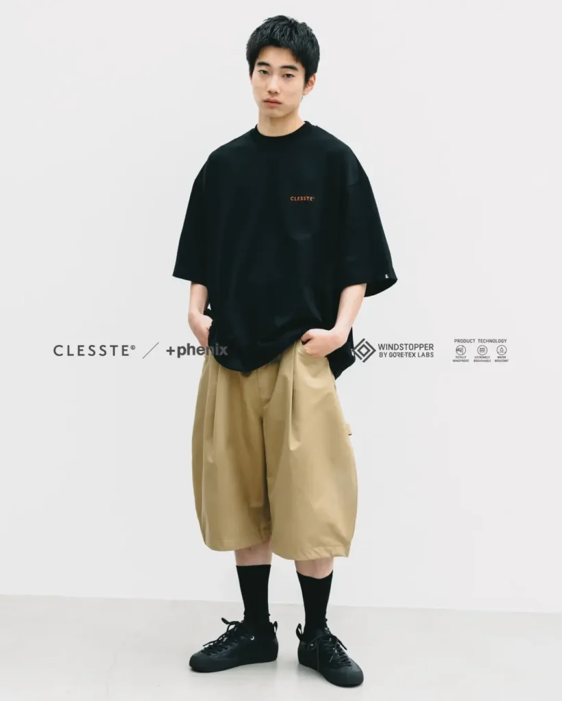Full-body front view of a model wearing a loose black oversized Tee with khaki balloon-fit technical shorts, black crew socks, and black shoe in a clean studio set