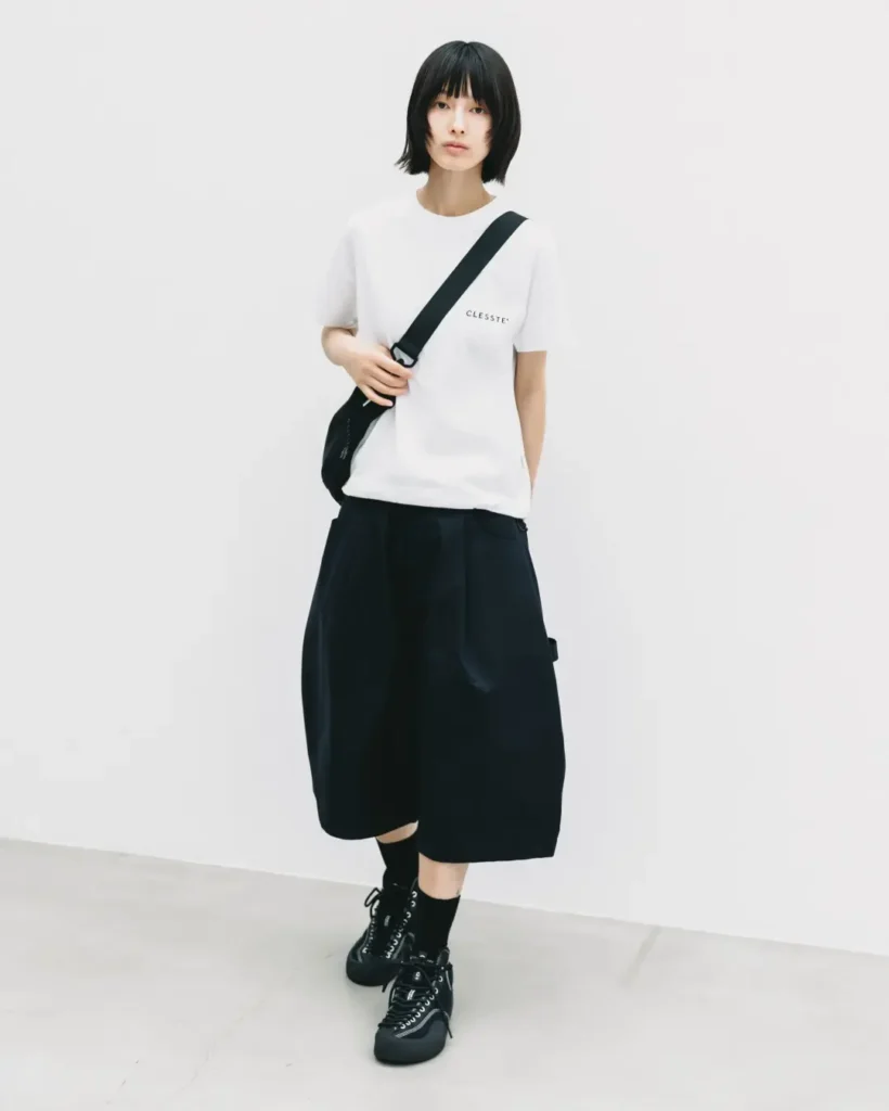 Full-body front view of a model wearing a white T-shirt with black oversized balloon shorts, a black crossbody shoulder bag, black socks, and black lace-up shoe against a white background