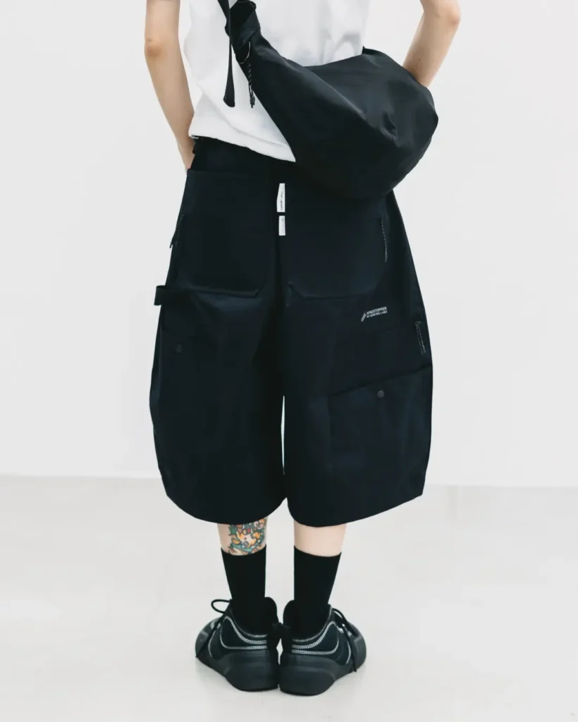 Rear close-up of black technical balloon shorts featuring oversized cargo pockets, zippered compartments, structured paneling, and a black crossbody bag worn over a white tee
