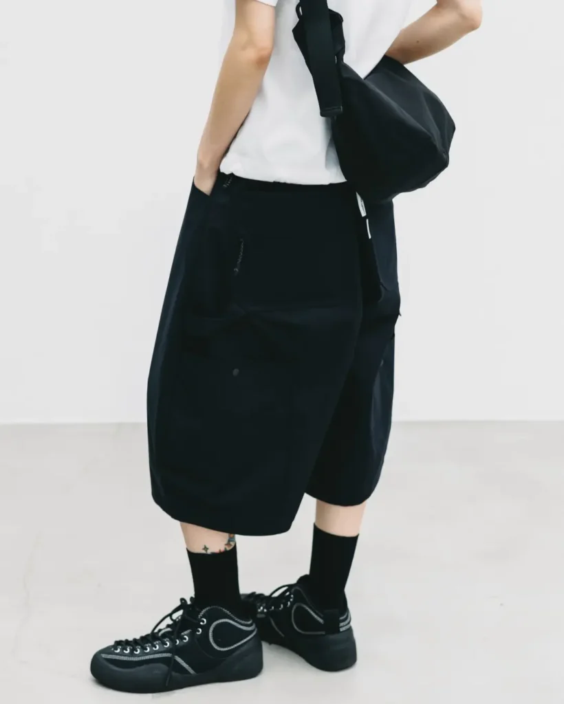 Three-quarter side view highlighting black oversized technical balloon shorts with utility pocket construction, paired with a white T-shirt, black socks, and black low-profile shoe