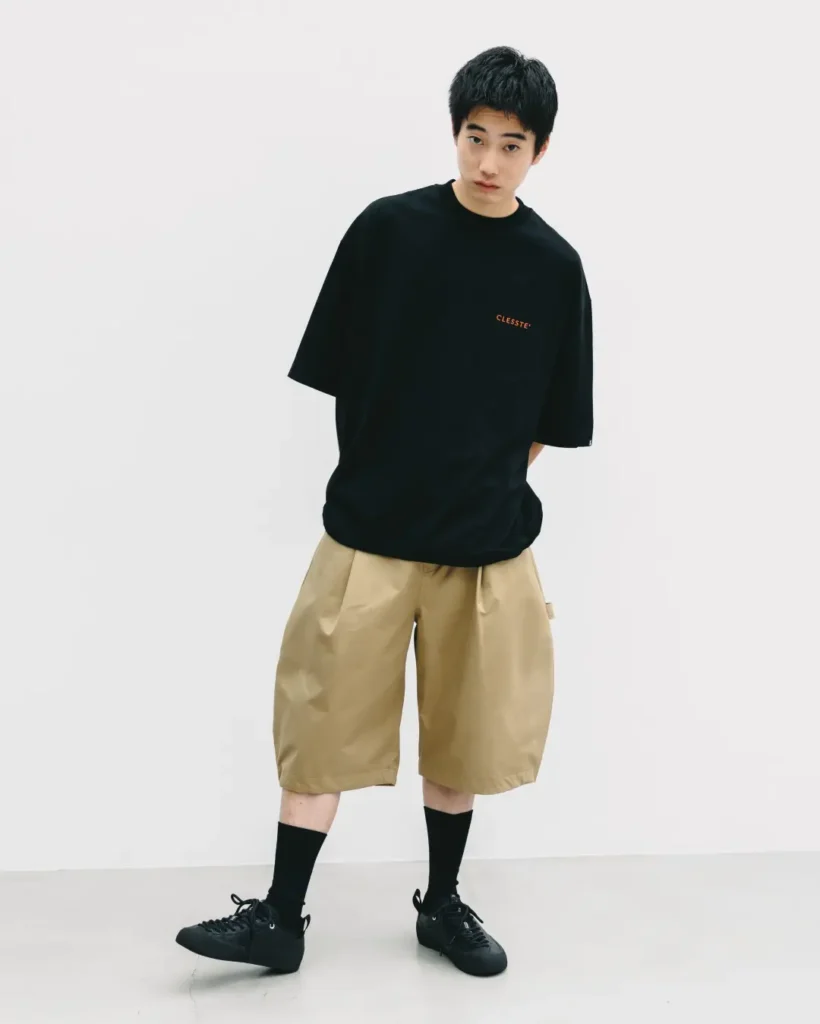 Full-body portrait of a model wearing an oversized black T-shirt with khaki balloon-fit technical shorts, black crew socks, and black shoe in a minimalist studio