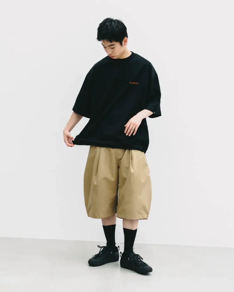 Full-body editorial image of a model adjusting an oversized black T-shirt while wearing voluminous khaki technical shorts with a relaxed wide-leg silhouette and black shoe