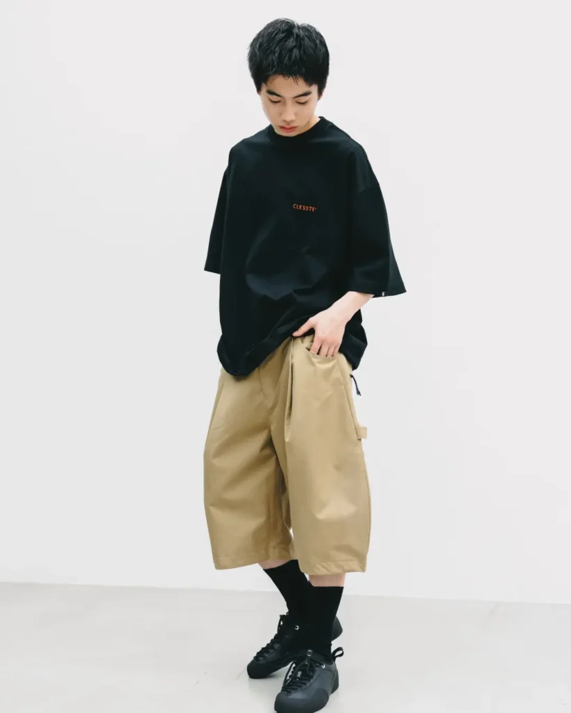 Front three-quarter view of a model styling an oversized black T-shirt with khaki balloon shorts featuring a roomy technical fit, black socks, and black lace-up shoe