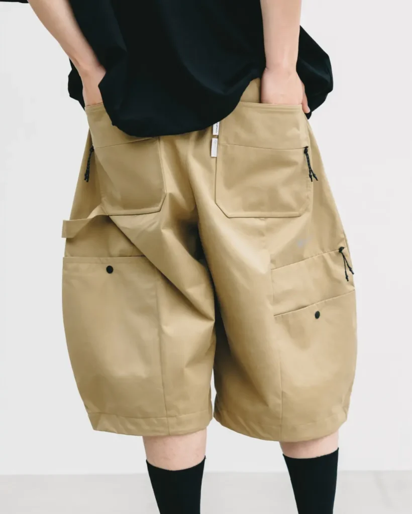 Rear close-up of khaki oversized utility shorts highlighting large cargo pockets, zip details, reinforced seams, and technical outdoor-inspired design beneath a black oversized