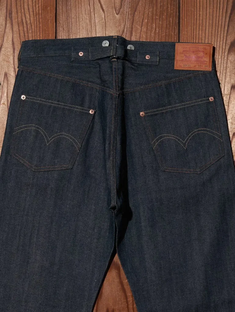 Rear upper section of Levi's 501 XX jeans showcasing the leather Two Horse patch, signature arcuate back pocket stitching, and clean raw indigo denim fin