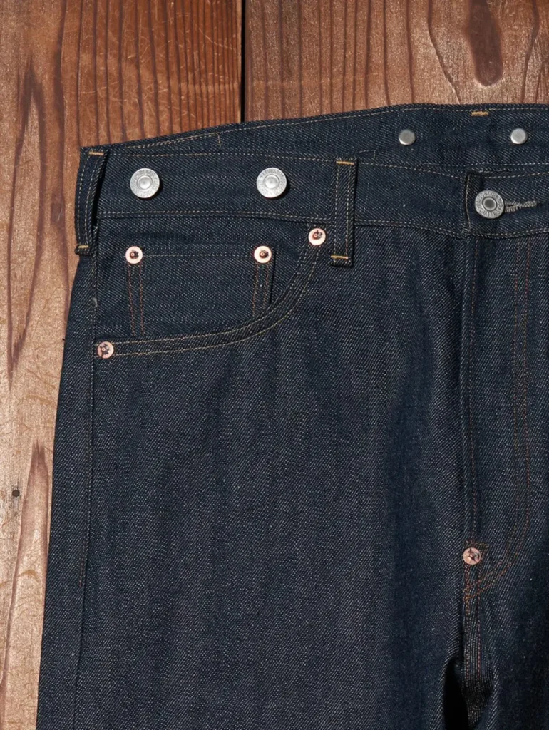 Close-up of the front left pocket area displaying copper rivets, reinforced coin pocket, belt loop, button fly waistband, and textured raw selvedge denim