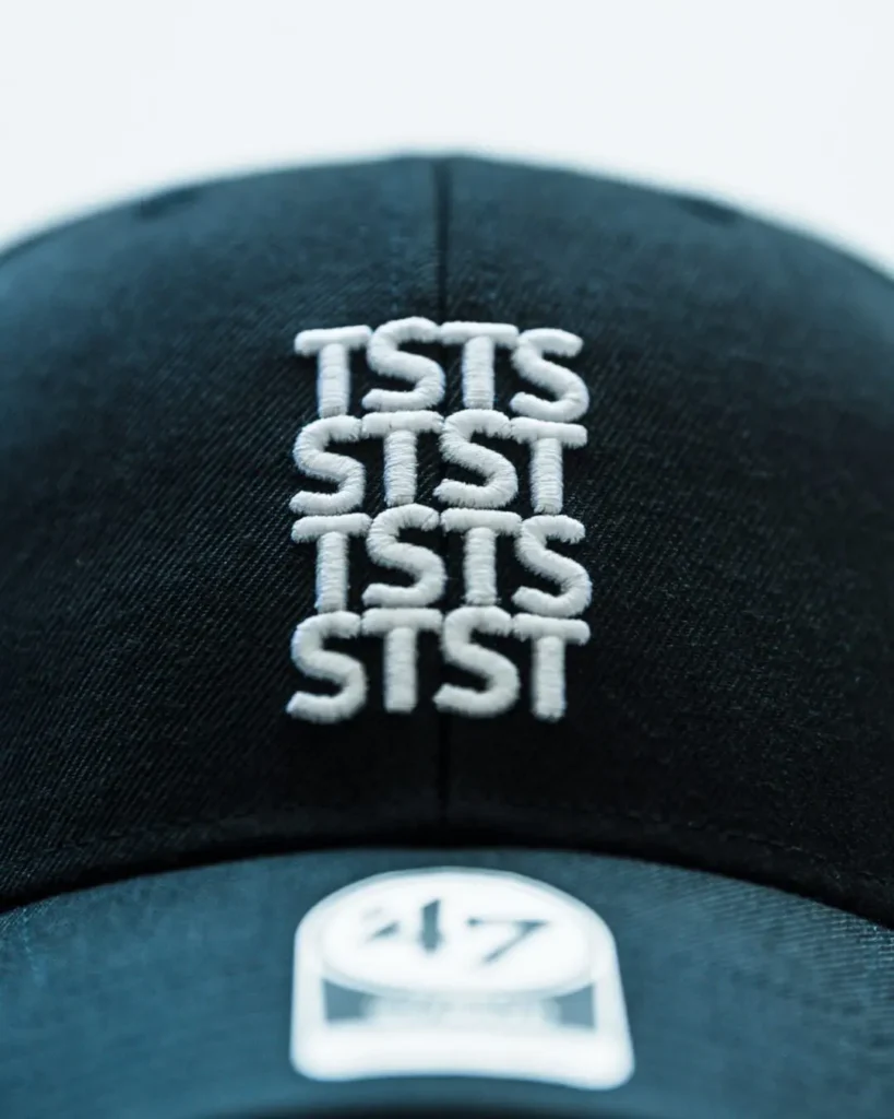 Close-up of the front panel highlighting the raised white TSTS embroidered logo arranged in a stacked grid pattern on the black cotton twill crown above the curved brim