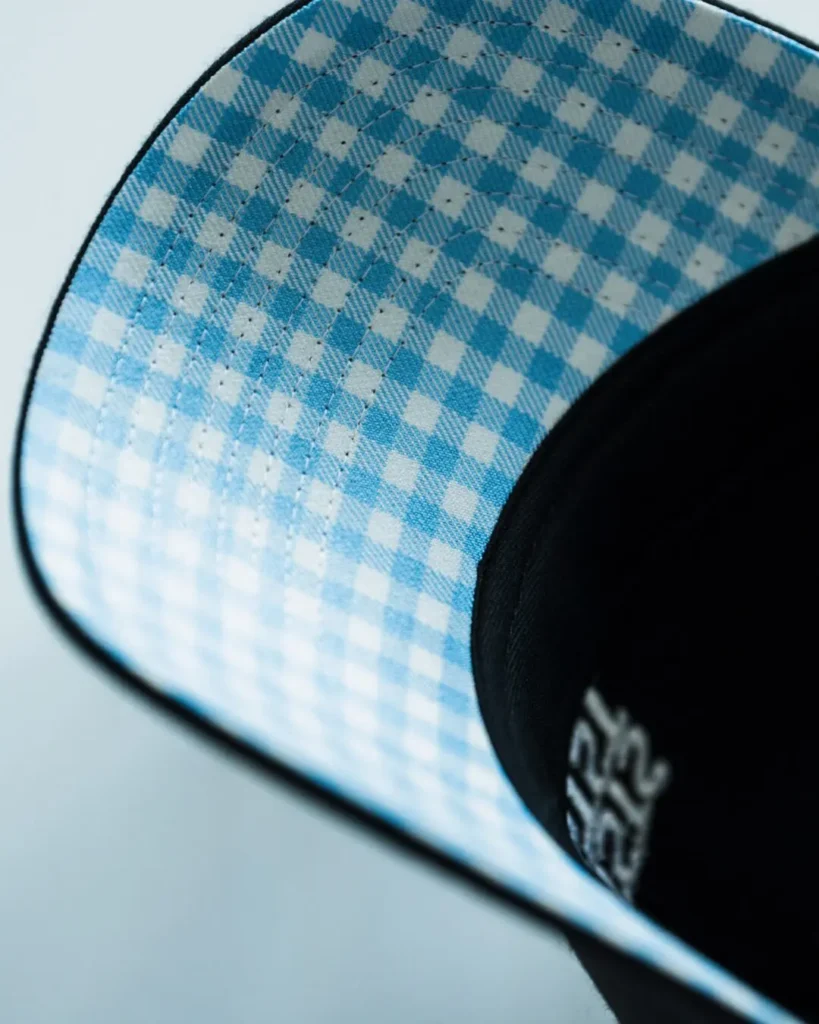 Detailed view of the underside of the cap's curved brim featuring a light blue and white gingham check pattern with concentric rows of stitching and black edge bind