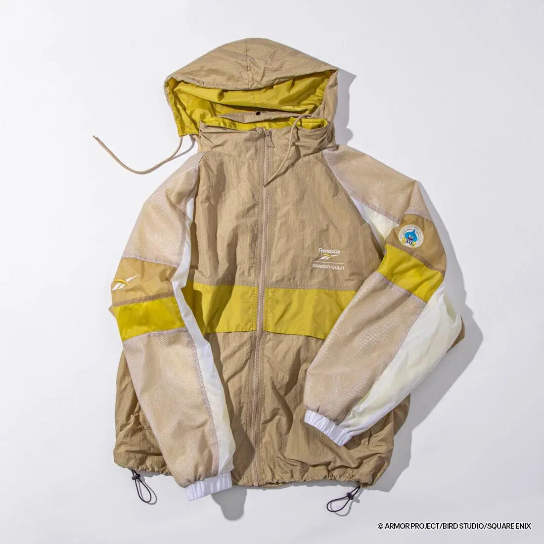 Beige and yellow Reebok × Dragon Quest hooded windbreaker featuring lightweight crinkled fabric, breathable mesh sleeves, a full-zip front, collaborative chest branding, and a Dragon Quest-themed sleeve patch.