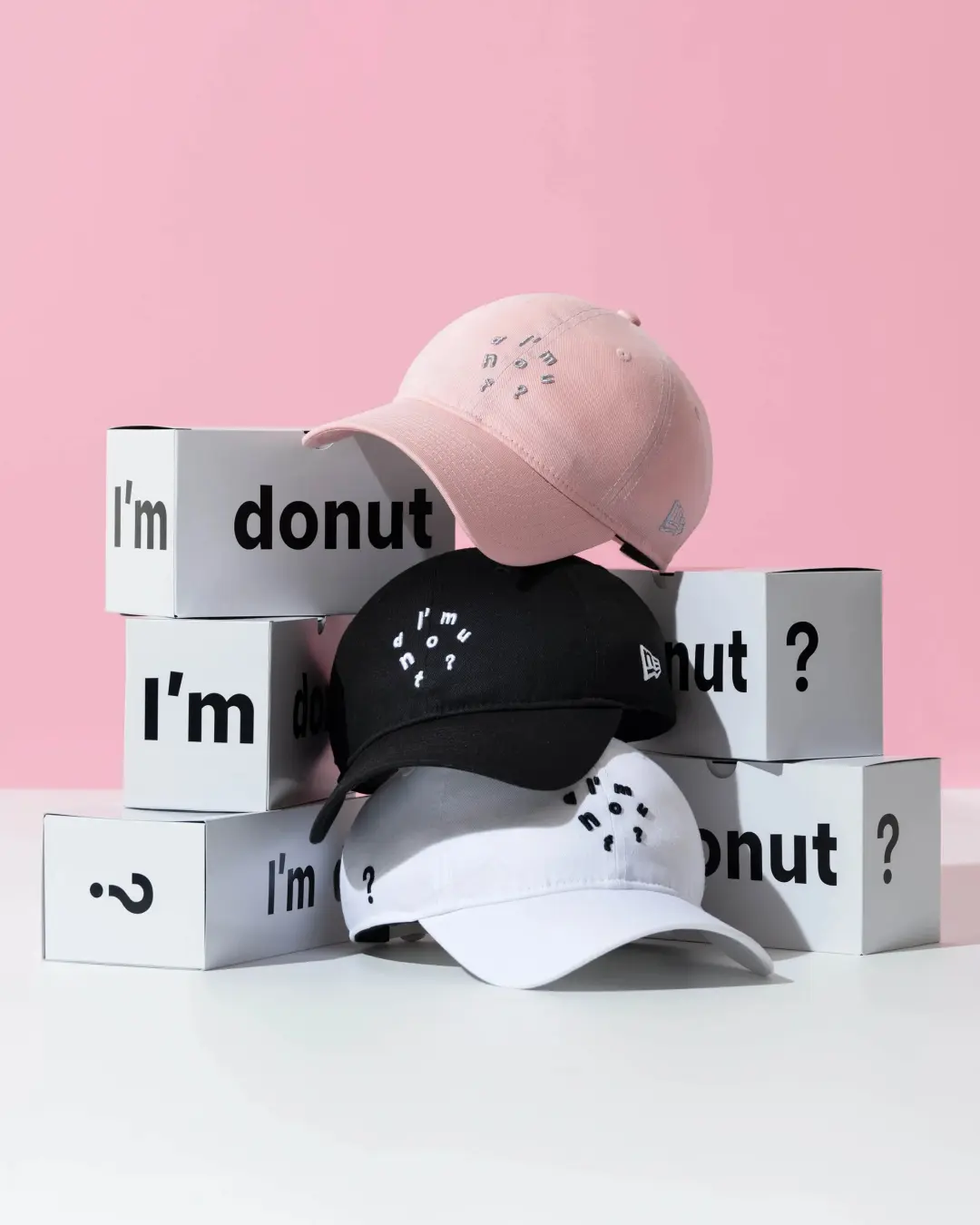 The complete I'm donut? × New Era 9TWENTY collaboration displayed in a clean studio composition featuring black, white, and pastel pink cotton caps arranged atop branded I'm donut? boxes. Each cap showcases the collection's signature scattered embroidered 'I'm donut?' lettering on the front, a curved visor, the embroidered New Era flag logo on the side, and minimalist detailing that reflects the playful identity of the cult Japanese doughnut brand. Set against a soft pink backdrop with balanced natural lighting, the image emphasizes the collaboration's understated aesthetic and collectible lifestyle appeal.