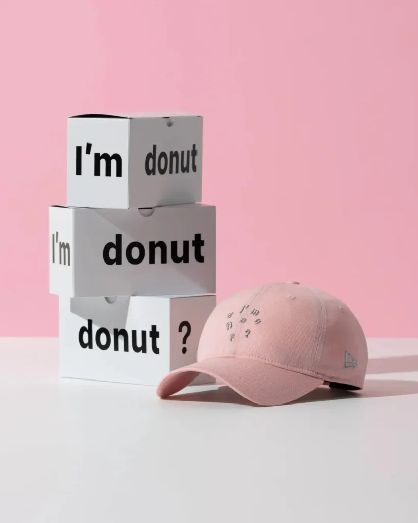 Pastel pink New Era "I'm Donut?" dad cap photographed beside stacked branded display boxes against a blush pink background, showcasing silver embroidered lettering and a matching side logo