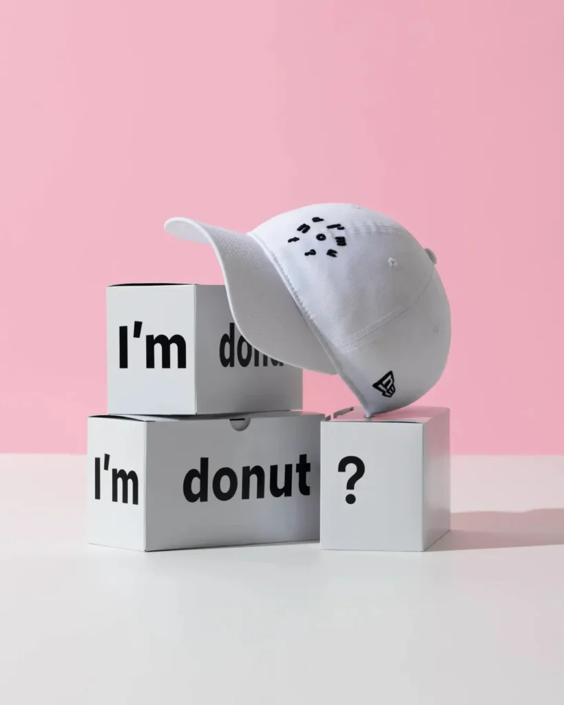 White New Era "I'm Donut?" dad cap balanced on branded white display boxes in a clean studio setup with a pastel pink backdrop, featuring black embroidered lettering and the signature New Era side logo