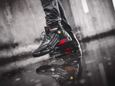 Air Jordan 14 "Last Shot" shoe worn outdoors on wet pavement, highlighting the black tumbled leather upper, glossy black midsole, red ribbed midfoot accent, and reflective puddle beneath the shoe