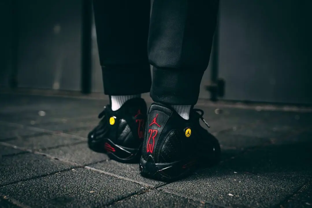 Rear on-foot view of the Air Jordan 14 'Last Shot' sneakers, showcasing the oversized red '23' heel embroidery, red Jumpman logo, Ferrari-inspired yellow shield badges, black tumbled leather construction, and white crew socks against a dark urban pavement.