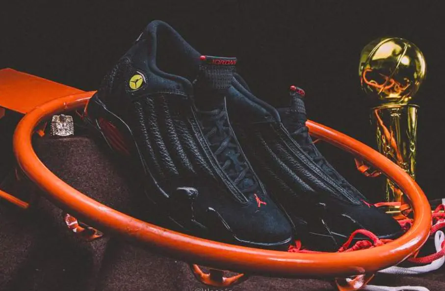 Air Jordan 14 'Last Shot' sneakers displayed inside an orange basketball rim with the Larry O'Brien Championship Trophy in the background, highlighting the black tumbled leather upper, red Jumpman accents, Ferrari-inspired yellow shield logo, and the silhouette's connection to Michael Jordan's final championship-winning shot.
