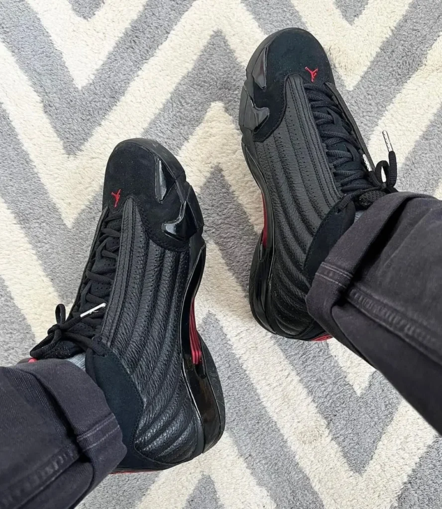 Top-down on-foot view of the Air Jordan 14 "Last Shot" revealing the stitched black leather vamp, black suede toe, red Jumpman embroidery on the toe box, and black pants over a patterned gray rug