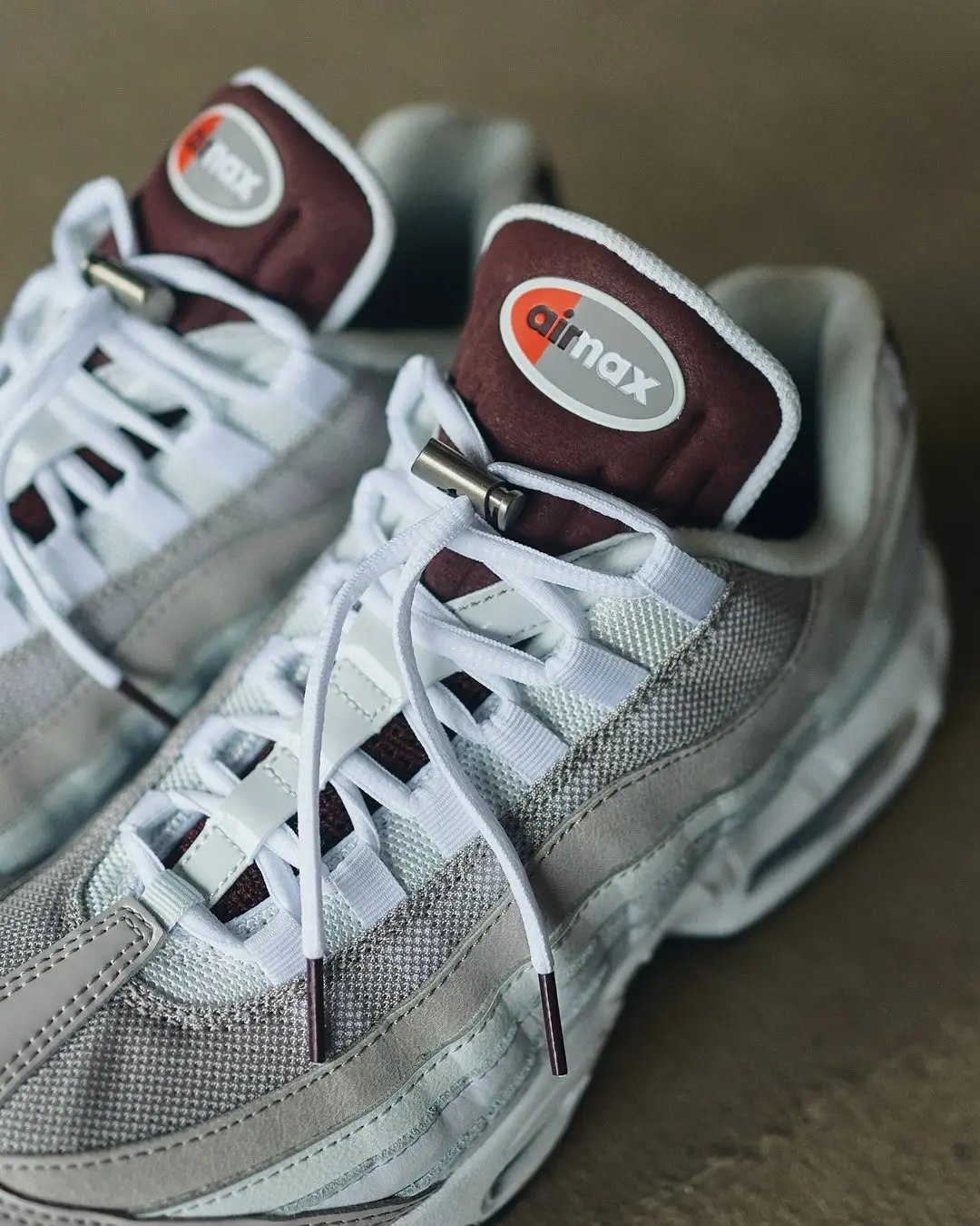 Close-up of the Nike Air Max 95 in a light grey and white colorway featuring layered mesh and suede panels, a padded brown tongue with the classic Air Max logo, white rope laces with a metallic lace lock, and visible Air cushioning.