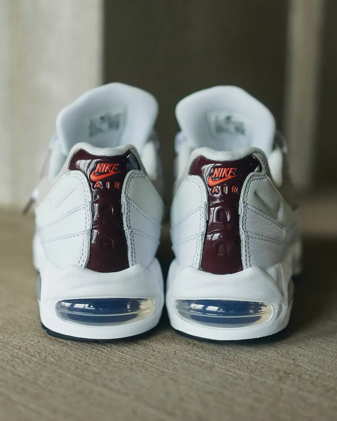 Rear view of the Nike Air Max 95 featuring glossy dark brown heel panels with bright orange Nike Air branding, white leather overlays, visible heel Air units, and a clean white midsole with black outsole accents.
