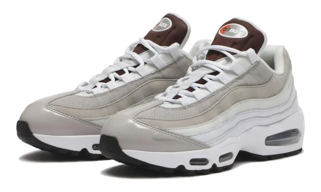 Three-quarter front view of the Nike Air Max 95 showcasing layered light grey suede and mesh uppers, white leather side panels, brown tongue branding, white laces, visible forefoot and heel Air units, and a black rubber outsole