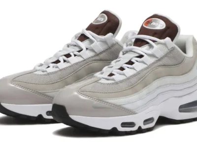 Three-quarter front view of the Nike Air Max 95 showcasing layered light grey suede and mesh uppers, white leather side panels, brown tongue branding, white laces, visible forefoot and heel Air units, and a black rubber outsole