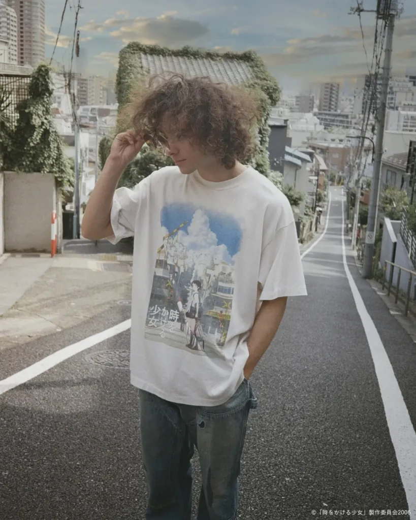 Curly-haired model adjusts their hair while wearing an oversized white graphic T-shirt featuring a vibrant illustrated Japanese city scene and anime character, paired with faded blue jean