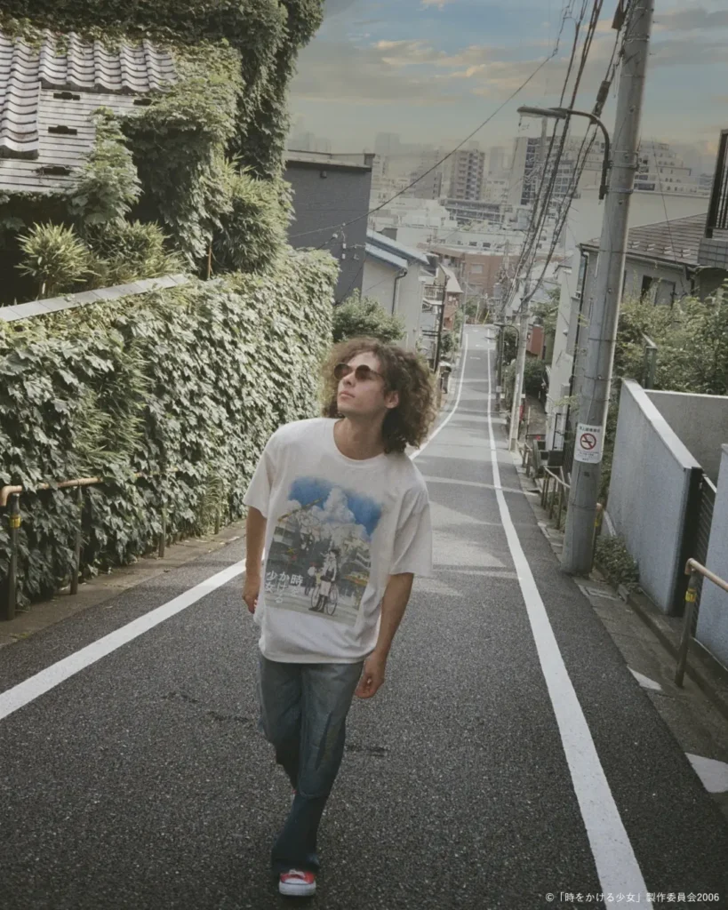 Lifestyle shot of a model in an oversized white anime graphic Tee walking up a sloped Tokyo street, wearing loose blue jeans and sunglasses for a casual streetwear look