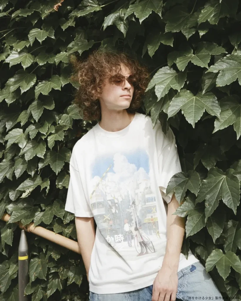 Model wearing an oversized white anime graphic T-shirt while posing against a wall covered in lush green ivy, highlighting the colorful illustrated front print and relaxed sil