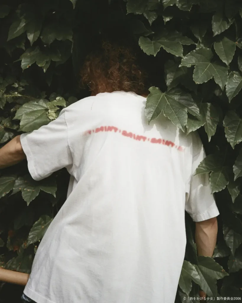 Rear view of an oversized white T-shirt featuring a blurred red graphic text print across the upper back as the model faces a dense wall of green ivy