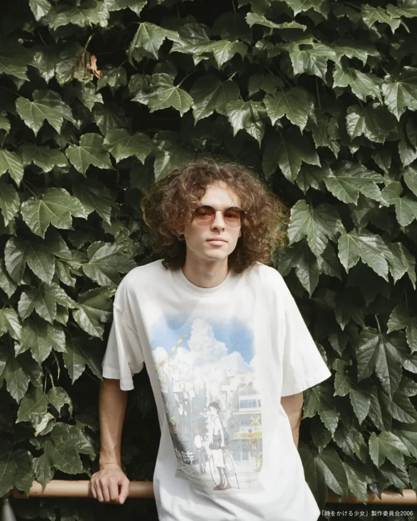 Portrait of a model leaning against a railing in front of lush ivy while wearing an oversized white graphic T-shirt with a detailed anime-inspired city illustration