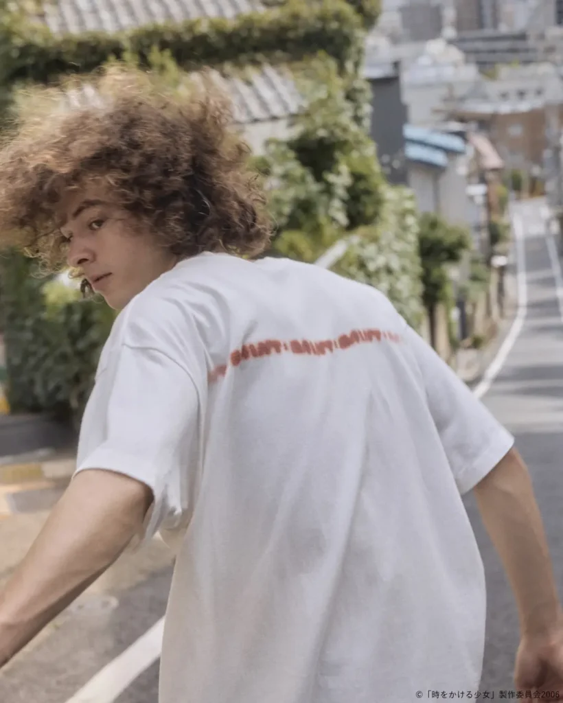 Rear three-quarter view of a model wearing an oversized white T-shirt with a blurred red text graphic across the upper back, captured on a sunlit residential street