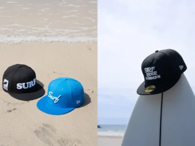 Two Surf Rider Foundation caps rest on a sandy beach beside the shoreline, while another black cap sits atop a white surfboard against a bright coastal sky
