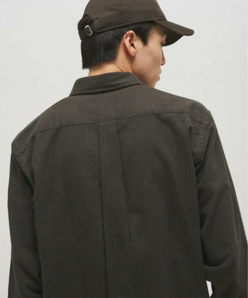 Rear view of a model wearing the dark charcoal Brooks Brothers British Check Shirt with a matching dark cap. The image highlights the back yoke, centered box pleat, relaxed shoulder construction, and subtle tonal check pattern across the brushed flannel fabric