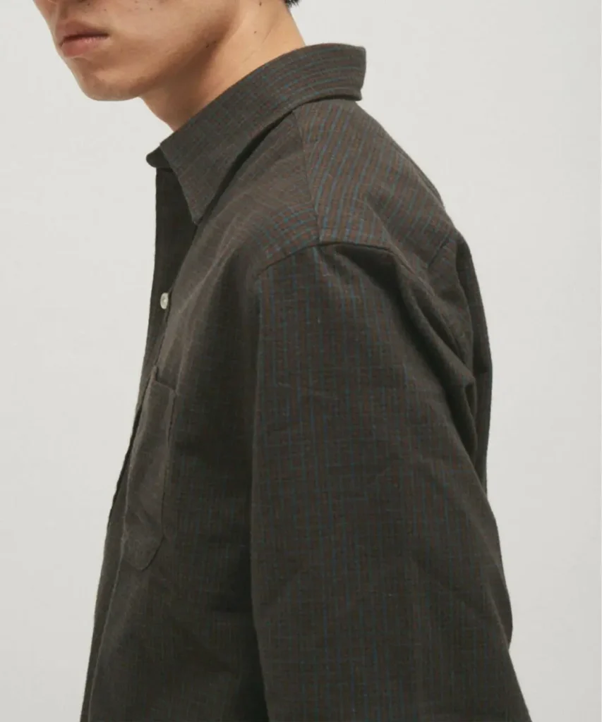 Side profile of a model wearing the charcoal Brooks Brothers check shirt. The close-up emphasizes the structured point collar, dropped shoulder, relaxed sleeve shape, chest pocket, and fine blue-and-brown check pattern woven into the dark brushed flannel