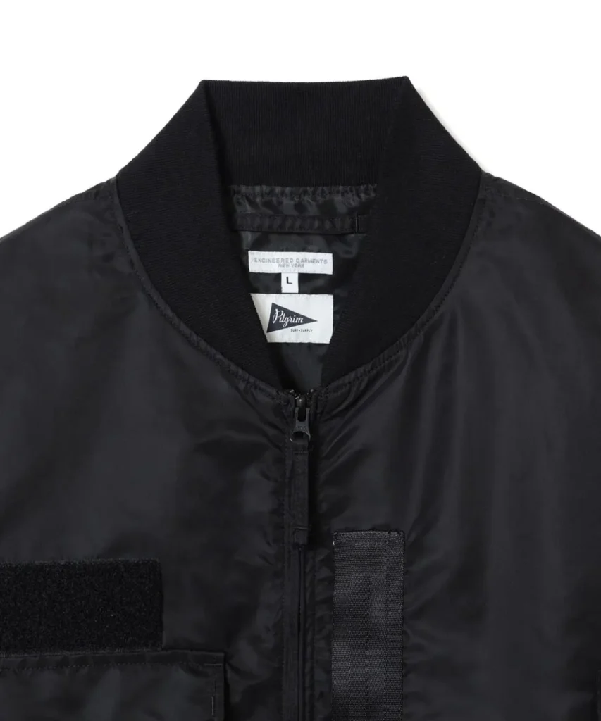 Close-up of the black bomber jacket's ribbed collar and zipper, showing the Pilgrim Surf + Supply and Engineered Garments labels, hook-and-loop chest panel, and reinforced webbing detail
