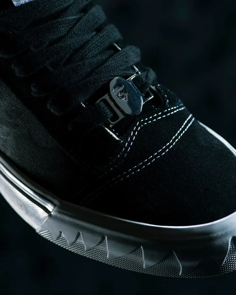 Detailed close-up of the forefoot featuring black suede, contrast white stitching, thick black laces, glossy metallic BAPE lace dubrae, and the jagged sculpted foxing wrapping the toe