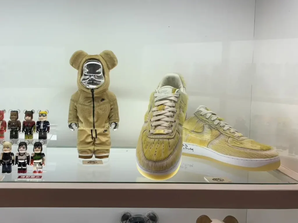 Display case featuring a tan Nike-themed Bearbrick-style figure with a reflective chrome face, flanked by two oversized fuzzy Nike Air Force 1 display shoes in a pale yellow colorway. Smaller collectible figures line the left side of the shelf inside a brightly lit exhibition case