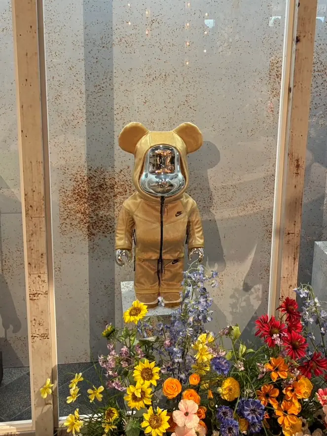 A gold Nike-themed BE@RBRICK figure with a mirrored chrome face stands inside a minimalist glass display. The collectible wears a matching gold hooded tracksuit featuring subtle Nike branding, while a vibrant arrangement of yellow, orange, blue, purple, and red flowers fills the foreground, contrasting with the clean exhibition backdrop.