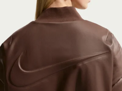 Rear close-up of a dark brown oversized Nike leather bomber jacket featuring an oversized embossed Swoosh spanning across the upper back. The model wears black wraparound sunglasses, silver hoop earrings, and straight blonde hair against a clean studio backdrop