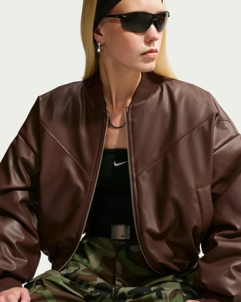 Front three-quarter view of a model wearing a dark brown oversized Nike leather bomber jacket with a zip-front closure, black Nike bandeau top, camouflage cargo pants, black headband, wraparound sunglasses, silver jewelry, and a metallic Nike belt in a minimalist studio set