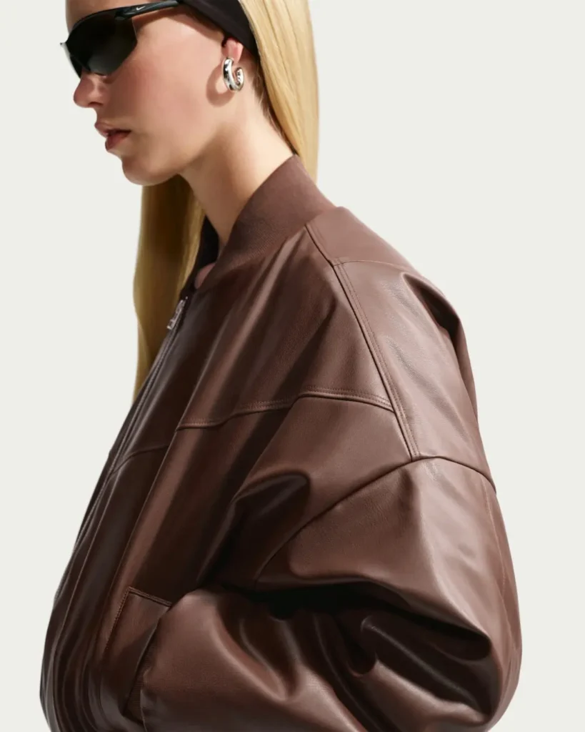 Side profile of the dark brown Nike leather bomber jacket highlighting its oversized silhouette, dropped shoulders, paneled construction, rib-knit collar, and smooth leather finish. The model wears black sport sunglasses and silver hoop earring