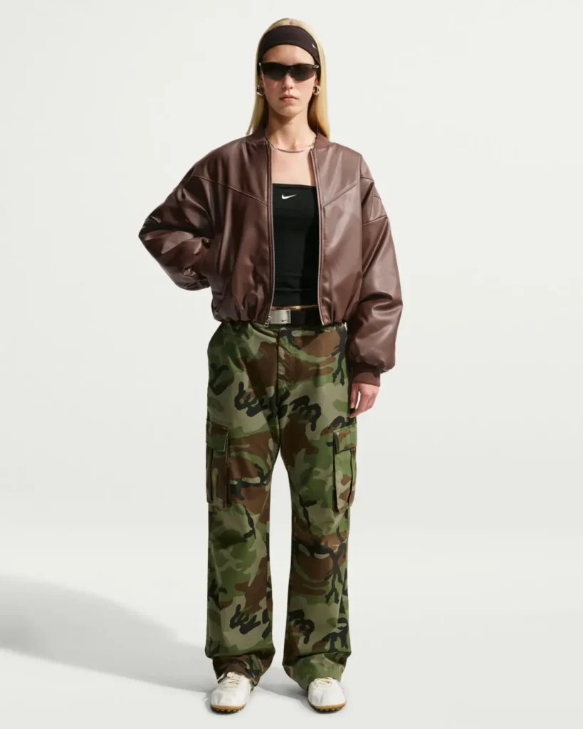 Full-body front view of the Nike leather bomber jacket styled with a black Nike bandeau top, woodland camouflage cargo pants, white sneakers, black headband, silver necklace, hoop earrings, and wraparound sunglasses in a neutral studio environment