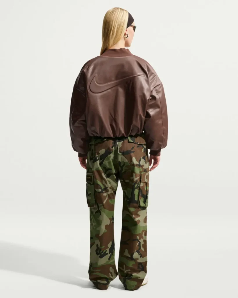 Rear full-body view of the oversized dark brown Nike leather bomber jacket showcasing the large embossed Swoosh across the back, ribbed hem and collar, voluminous sleeves, and camouflage cargo pants styled with white sneakers against a clean studio background