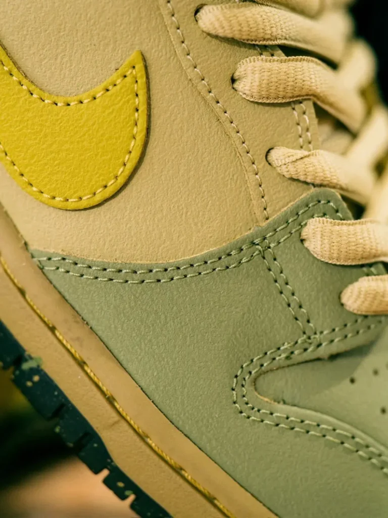 Macro shot highlighting the textured yellow leather Swoosh, cream quarter panel, sage green mudguard, and cream flat lace